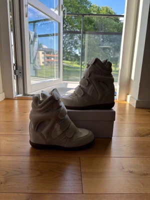 Isabel Marant vita high top sneakers - Snygga vita high top sneakers från Isabel Marant med chunky siluett och flera kardborreband. Skorna har en blandning av mocka och textil, samt en beige sula som ger en cool kontrast. Perfekta för dig som vill ha en statement sneaker med unik design.