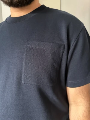 Mörkblå Louis Vuitton t-shirt med bröstficka - Snygg mörkblå t-shirt med diskret bröstficka och klassisk rund hals. T-shirten har diskret Louis Vuitton monogram på bröstfickan samt hela ryggen. Den är i storlek L men passar även de som bär M lika väl. T-shirten är slut i lager, då den är en del av deras nya kollektion. Materialet känns mjukt och skönt mot huden. Säljer den då jag inte har någon vidare användning för den, köpt i LV butiken i Milano. Kvitto, låda samt påse medföljer. Använd ett fåtal gånger (ca 2-3 gånger). Pris kan diskuteras