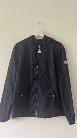 Mörkblå Moncler cardigan - Ej använd mörkblå vindjacka från Moncler med huva och dragsko. Jackan har dragkedja framtill, två fickor med tryckknappar och Moncler-logga på ärmen. Huvan har en vit och röd rand som detalj. Perfekt för blåsiga dagar. Storlek S, funkar för M också. Inga returer! Det går även att sänka priset, bara att meddela!