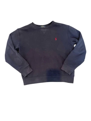 Mörkblå sweatshirt från Polo Ralph Lauren - Klassisk mörkblå sweatshirt från Polo Ralph Lauren med rund halsringning och röd broderad logga på bröstet. Tröjan har ribbade muddar vid ärmslut och nederkant, perfekt för en avslappnad och stilren look. Storlek 14-16 år som  motsvarar ca xs