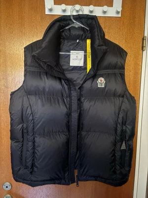 Svart dunväst från Moncler - Snygg svart dunväst från Moncler med hög krage och dragkedja framtill. Västen har två framfickor med dragkedja och Moncler-logga på bröstet. Klassisk puffer-design i glansigt material som ger en sportig och trendig look.