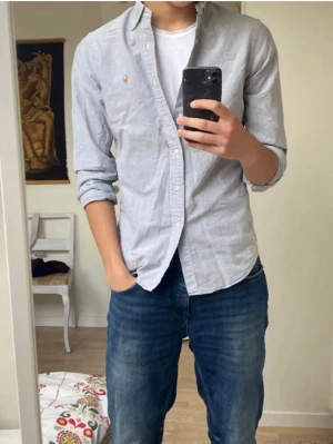 Ljusblå skjorta från Ralph Lauren - Klassisk ljusblå skjorta från Ralph Lauren med knappstängning och broderad logga på bröstet. Skjortan har lång ärm och normal passform, tillverkad i mjuk bomull. Perfekt till jeans eller chinos för en clean och stilren look. Små slitningar vi kragen men knappt något som märks.