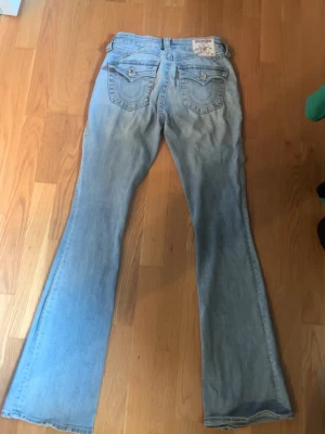 Ljusblå bootcut jeans True Religion - Säljer ett par ljusblå bootcut jeans från True Religion med klassiska kontrastsömmar och broderade bakfickor. Jeansen har låg midja och är tillverkade i denim med lätt tvättad look. Perfekta för dig som gillar retrostil och vill sticka ut.