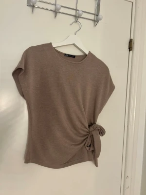 Beige topp med knyt från Zara - Säljer en beige topp från Zara med rund halsringning och korta ärmar. Toppen har en snygg knyt-detalj i sidan som ger en cool twist till looken. Perfekt att styla med jeans eller kjol för en trendig outfit.