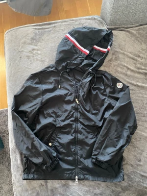 Mobcler windbreaker - Snygg svart vindjacka från Moncler med huva och dragsko. Jackan har dragkedja framtill, två fickor med knapp och Moncler-logga på ärmen. Huvan har en röd, vit och blå rand som ger en sportig vibe. Tillverkad i lätt och glansigt material. Storlek S, inga skador eller hål