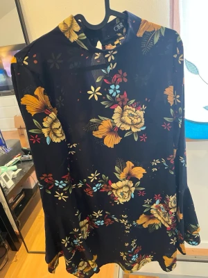 Mörkblå blommig klänning från CBR - Mörkblå klänning från CBR med färgglatt blommönster i gult, rött, blått och vitt. Klänningen har lång ärm, hög hals med knappdetalj bak och en lös, rak passform. Tillverkad i ett lätt och mjukt material som ger en skön känsla.