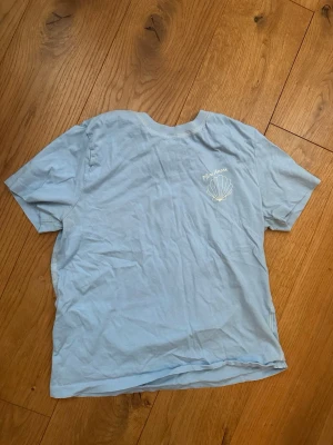 Ljusblå T-shirt  - Ljusblå T-shirt med en vit snäcka i storlek S