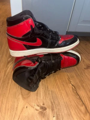 Air Jordan 1 High Bred Svart/Röd - Säljer ett par ikoniska Air Jordan 1 High sneakers i svart och rött med vita sulor. Skorna har klassisk Nike Swoosh på sidan, högt skaft och är tillverkade i glansigt läder. Perfekta för dig som gillar streetwear och vill sticka ut med en tidlös modell.