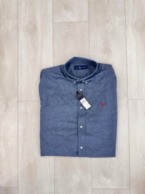 Blå skjorta från Ralph Lauren - Klassisk blå skjorta från Ralph Lauren med button-down krage och vita knappar. Skjortan har en liten röd broderad logga på bröstet och är tillverkad i bomull. Perfekt för en stilren och avslappnad look.
