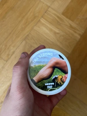 Uffe dosa  - Säljer en rund snusdosa från MUF med texten 'EXTRA STRONG' på locket. Dosan är vit med ett färgglatt motiv på ovansidan som visar ett ben och gräs. Praktisk att ha med sig och enkel att använda.