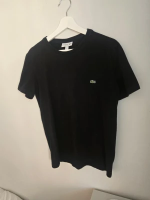 Svart t-shirt från Lacoste - Klassisk svart t-shirt från Lacoste med rund hals och den ikoniska gröna krokodilloggan broderad på bröstet. Tillverkad i mjuk bomull och har en enkel, stilren design som passar till det mesta.