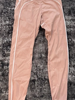 Beige set från Alo Yoga - Snygg beige sport-bh från Alo Yoga med vita detaljer och bred resår under bysten. Bh:n har en mjuk och stretchig passform med vadderade kupor och är perfekt för yoga eller träning. Materialet är slätt och bekvämt mot huden. Byxorna kommer även med.