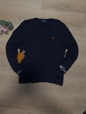 Mörkblå kabelstickad tröja Polo Ralph Lauren - Mörkblå kabelstickad tröja från Polo Ralph Lauren med klassisk orange logga broderad på bröstet. Tröjan har rund halsringning, ribbade muddar och nederkant. Perfekt för dig som gillar stilrena och tidlösa plagg.