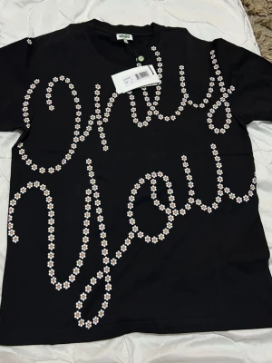 Svart Kenzo t-shirt med blommönster - Snygg svart t-shirt från Kenzo med texten 'Only You' skriven i små vita blommor över hela framsidan. Klassisk rund hals och korta ärmar. Tillverkad i mjuk bomull som känns skön mot huden. Perfekt för dig som vill sticka ut med en cool och lekfull design.