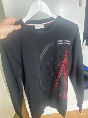 Svart sweatshirt från Moncler - Svart sweatshirt från Moncler med stort broderat logotypmönster i svart och rött på framsidan. Tröjan har rund halsringning, långa ärmar och en liten Moncler-patch på ena ärmen. Tillverkad i mjukt bomullsmaterial för en skön känsla.