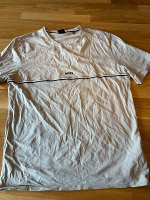 Biege t-shirt från BOSS med tryck - Snygg vit t-shirt från BOSS med svart tryckt logga på bröstet och en tunn svart linje tvärs över framsidan. Klassisk rund halsringning och korta ärmar. Tillverkad i mjuk bomull som känns skön mot huden. Perfekt till jeans eller shorts.aldrig använd bara legat längst nere i lådan.
