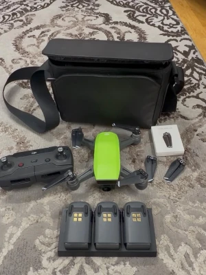 DJI Spark drönare med tillbehör - DJI Spark drönare i grön färg med svart fjärrkontroll, tre batterier med laddningsstation, extra propellrar och en svart väska. Kompakt design i plast och metall, perfekt för flygfoto och video. Allt du behöver för att komma igång med drönarflygning.