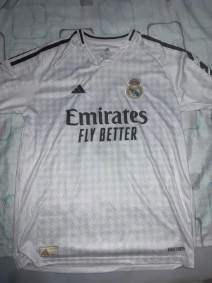 Real Madrid Mbappé matchtröja vit - Snygg vit Real Madrid matchtröja från Adidas med Mbappé och nummer 9 på ryggen. Tröjan har svarta detaljer, klubbmärke på bröstet och sponsortryck framtill. Tillverkad i lätt och ventilerande material med HEAT.RDY-teknologi. Står XXL men skulle säga att den är XL