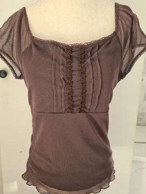 Brun mesh topp från KappAhl - Söt brun mesh topp från KappAhl med korta transparenta ärmar och dekorativa spetsdetaljer framtill. Toppen har en lätt figurnära passform och vågig kant nedtill. Perfekt för dig som gillar romantiska detaljer och mjuka material.