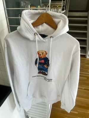 Vit hoodie Polo Ralph Lauren björn - Vit hoodie från Polo Ralph Lauren med den ikoniska björnen på bröstet, klädd i blå tröja och jeans. Hoodien har dragsko i huvan och en stor magficka. Tillverkad i mjukt bomullsmaterial och har en avslappnad passform. Storlek M