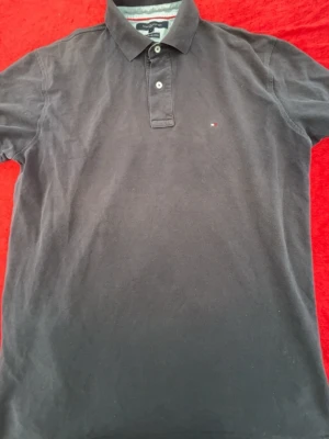 Svart pikétröja Tommy Hilfiger - Klassisk svart pikétröja från Tommy Hilfiger med liten logga på bröstet och två knappar vid kragen. Tillverkad i mjuk bomull och har korta ärmar. Perfekt för en clean och stilren look.