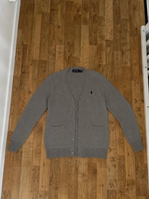 Grå cardigan från Polo Ralph Lauren - Klassisk grå kofta med V-ringning från Polo Ralph Lauren. Tröjan har två framfickor, ribbade muddar och knäppning framtill. Liten mörkblå logga broderad på bröstet. Perfekt för lager på lager och stilren look. L från början men tvättat fel så den passar som M