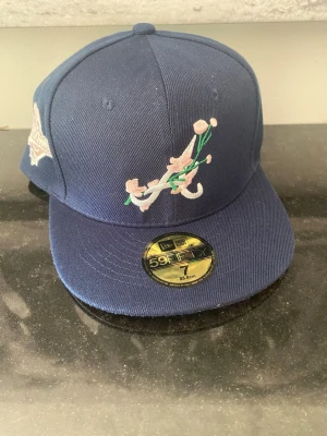 Blå New Era 59Fifty Atlanta keps - Säljer en blå fitted keps från New Era, modell 59Fifty, med Atlanta Braves-logga framtill dekorerad med broderade blommor i rosa och grönt. Klassisk rak skärm och broderad logga på sidan. Perfekt för dig som gillar streetwear och sportig stil.