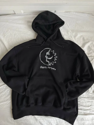 Svart hoodie District FortySix - Svart hoodie från District FortySix med stor vit grafisk print på bröstet som föreställer ett ansikte i profil och texten 'district forty six'. Hoodien har huva med dragsko och en stor magficka. Tillverkad i mjukt material, perfekt för en avslappnad stil.