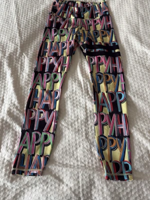 Färgglada leggings med HAPPY-tryck - Säljer ett par riktigt färgglada leggings med ordet 'HAPPY' i stora bokstäver över hela plagget. Mönstret går i flera färger som gul, blå, rosa och svart. De har en tajt passform och är tillverkade i ett stretchigt material som känns bekvämt. Endast använda ett fåtal gånger. 