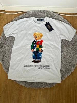 Vit Polo Bear t-shirt  - Vit t-shirt med ikoniskt Polo Bear-tryck på framsidan. Björnen har färgglada kläder och jeans, och under motivet står det 'Polo Bear by Ralph Lauren'. Klassisk rund hals och korta ärmar.