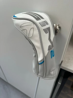 TaylorMade SIM2 Max golfklubba trä trea  - Golfklubba TaylorMade SIM2 Max med svart, vit och blå design. Klubban har ett modernt utseende med tydliga logotyper och detaljer i gult och blått. Medföljer ett vitt och grått headcover med TaylorMade-logga och blå accent.