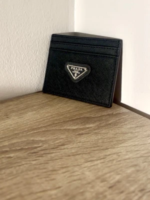 Plånbok - Snyggt svart kortfodral i äkta läder med klassisk trekantig metallogo framtill. Fodralet har flera kortfack och stilren design som passar perfekt i fickan. Lyxig känsla och tidlös look för dig som gillar exklusiva accessoarer.