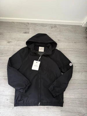 Svart vindjacka Moncler storlek M - Snygg svart vindjacka från Moncler med huva och dragkedja framtill. Jackan har elastiska muddar vid ärmar och nederkant samt en ficka på ärmen med Moncler-logga. Tillverkad i lätt och slitstarkt material, perfekt för blåsiga dagar. Kom dm för frågor, bud eller byten