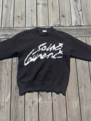 Saint Laurent tröja - Säljer en svart stickad tröja från Saint Laurent med vit grafisk text på bröstet. Tröjan har rund halsringning och ribbade muddar. Materialet ser mjukt och fluffigt ut, perfekt för dig som gillar streetwear och exklusiva märken. Jag är 176 och den sitter perfekt på mig! Hojta till om det är något!