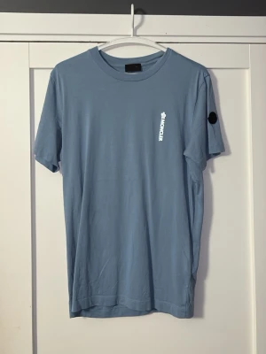 Blå Moncler t-shirt - Snygg ljusblå t-shirt från Moncler med vit logga tryckt på bröstet och ett svart Moncler-märke på ärmen. Klassisk rund halsringning och korta ärmar. Tillverkad i mjuk bomull för skön känsla och stilren look.