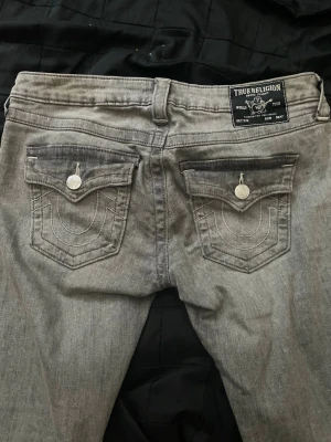 Gråa True Religion jeans - Säljer ett par gråa jeans från True Religion med ikoniska bakfickor och silverfärgade knappar. Jeansen har låg midja, utsvängda och fickor där bak. Jättesnygga på men säljer då jag ej använder de så mycket. Nypris var runt 2200. Storlek 28 vilket motsvarar ca S