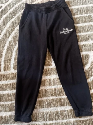 Svarta mjukisbyxor Peak Performance - Svarta mjukisbyxor från Peak Performance med tryckt logga på vänster sida. Byxorna har resår i midjan och vid bensluten samt två sidofickor. Perfekta för chill eller träning. Mjukt och bekvämt material.