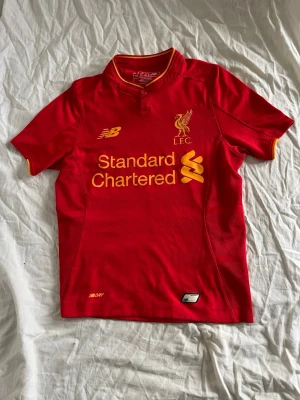 Liverpool FC röd matchtröja - Liverpool FC matchtröja i rött med gula detaljer och klubbmärke på bröstet. Tillverkad i lätt funktionsmaterial med NB Dry-teknologi. Kortärmad modell med knapp i halsen och tryckt sponsorlogga på framsidan.