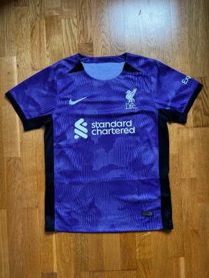 Lila Liverpool FC matchtröja Nike - Snygg lila Liverpool FC matchtröja från Nike med vita loggor och sponsortryck på bröstet. Tröjan har korta ärmar, svarta sidopaneler och ett subtilt mönster i tyget. Perfekt för fotboll eller träning.