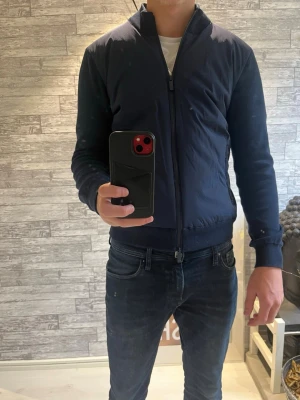 Mörkblå jacka från John Henric - Snygg mörkblå bomberjacka från John Henric med dragkedja framtill och två fickor med dragkedja. Jackan har ribbade muddar vid ärmslut och nederkant samt en minimalistisk design. Perfekt för dig som gillar stilrena plagg med modern känsla.