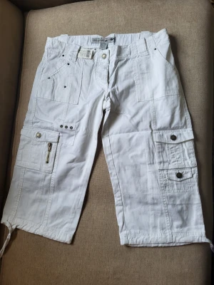 Vita cargoshorts med fickor Wet Seal Storlek Us 9 L - Säljer ett par vita cargoshorts med flera praktiska fickor, dragkedjedetalj och knappar. Shortsens längd går ner till knäna och de har snörning vid bensluten. Perfekta för en avslappnad och trendig stil i sommar.