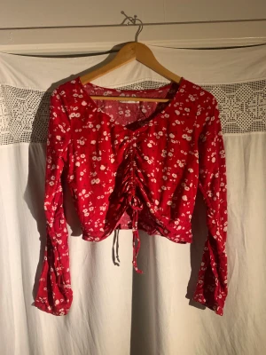  Croppad topp från Hollister - Väldigt söt topp från Hollister i röd med vita blommor. Långa ärmar och justerbara band längs med framsidan för att kunna justera längden/formen (se bild 1 & 2). Storlek S. Använd runt 1-2 ggr, mycket bra skick!