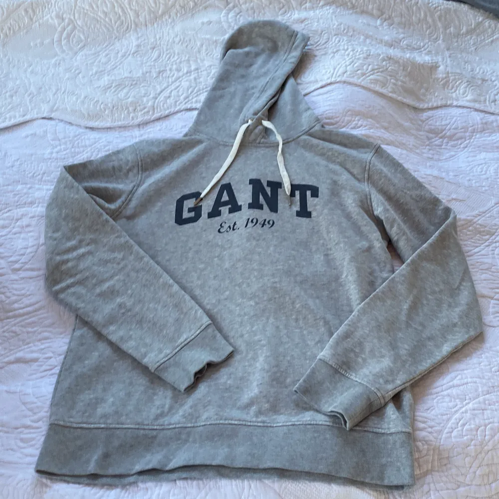 En grå lite tjock gant tröja med en mörkblå text där det står” GANT est 1949. Med två vita snören vid luvan . Tröjor & Koftor.