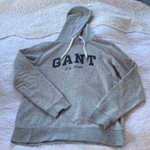 En grå lite tjock gant tröja med en mörkblå text där det står” GANT est 1949. Med två vita snören vid luvan 