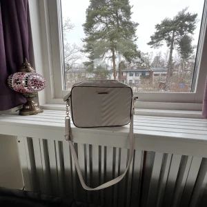 Väska  - En beige axelväska med detaljer i guld. Den har ett fack på insidan och är i hyfsat gott skick. 