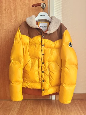 Gul dunjacka med fårskinnskrage - Blast-Off Sirio Basic Quilted Puffer Jacket  Gul dunjacka i unikt utförande. Uppvikbar fårskinnskrage och axlar i brun mocka.  Mycket bra skick.  Size 48, kan passa både S och M.  Spana gärna in mina andra annonser också!  DM vid frågor.