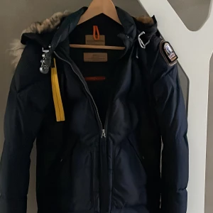 Parajumper long bear dam marinblå - Long Bear marinblå kappa med  ikoniska Parajumpers detaljer som patchen, cargo pocket och gult elastiskt band inspirerat av det som används av fallskärmsjägare.  Storlek S. Normal passform. Två fickor fram slits med dragkedja i sidorna. Muddar i ärmslut. 