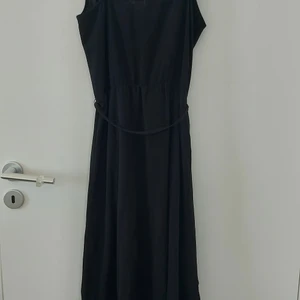 Svart klänning med detaljer  - Svart klänning med omlottkjol, volang detaljer samt ett knytband runt midjan. Finns även en liten knapp fram till vid urringning. Priset 