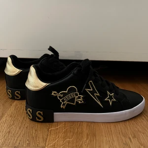 Guess sneakers  - Säljer mina jätte coola Guess sneakers i svart med  guld detaljer, säljer pga att de är för små. Sneakersarna är använda ett fåtal gånger och är i nyskick. På baksidan står Guess i guld och det är en stjärna, blixt och ett hjärta i guld på ena sidan på båda skorna. Skorna är i storlek 39.