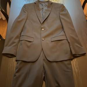 Svart kostym från Dressman, byxorna har pressveck och kavajen har två knappar. Storleken på byxorna är W27 och L30, kavajen är i storlek 44. Detta skulle motsvara xs